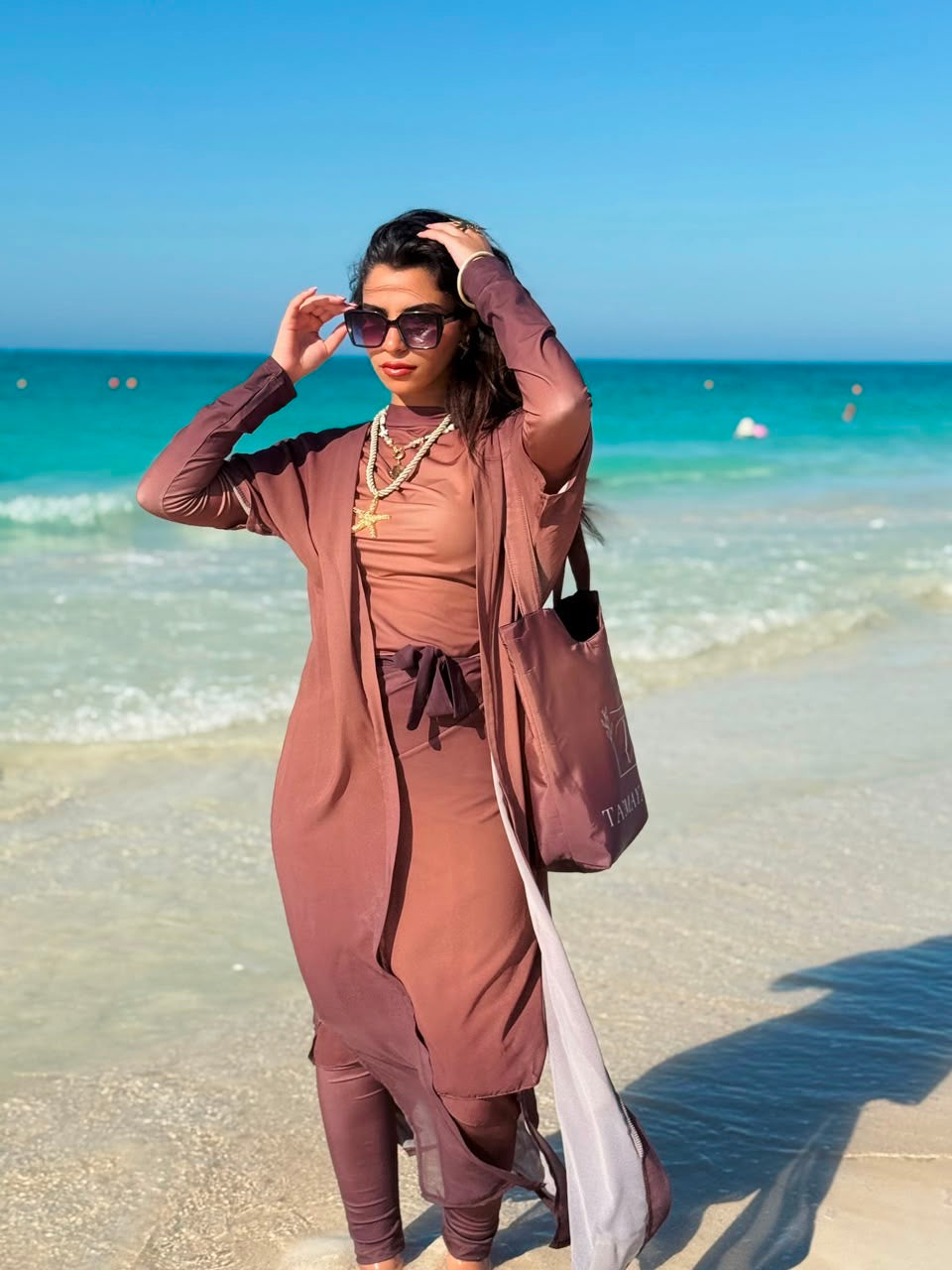 Ta’mayaa swimwear modest full coverage set in brown worn by woman on the beach with ocean background