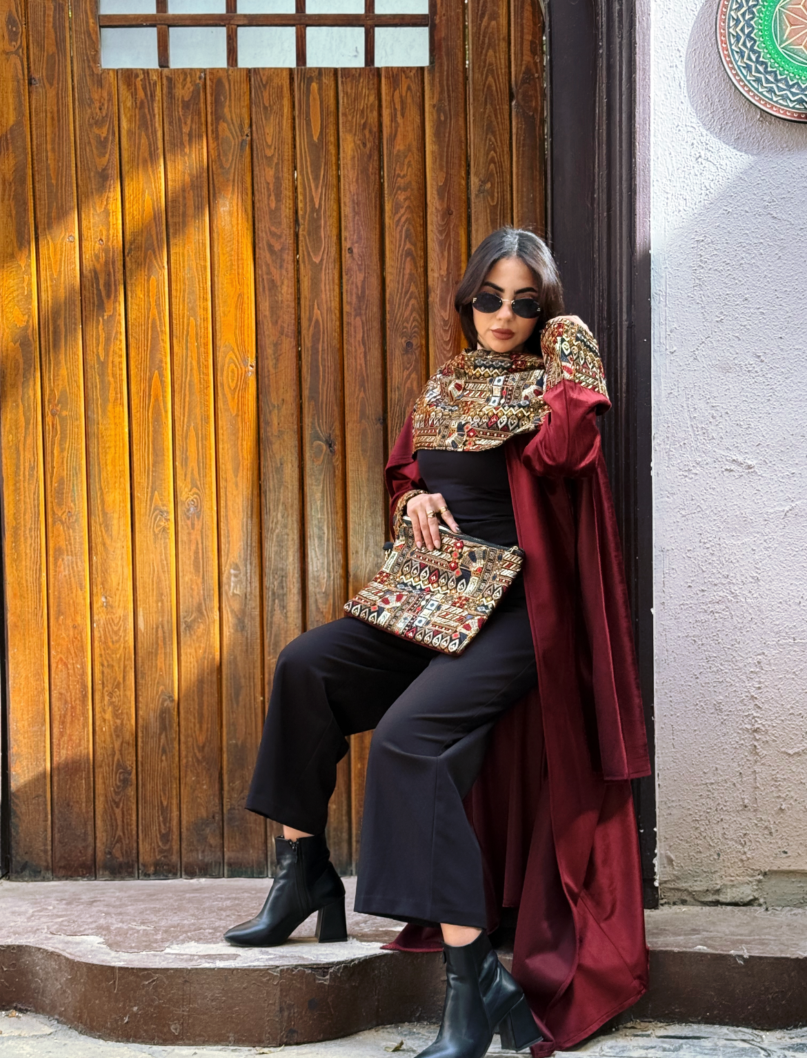 Woman wearing a Burgundy Elegance Embroidered Velvet Kaftan with geometric embroidery, accessorized with a clutch in a rustic setting.