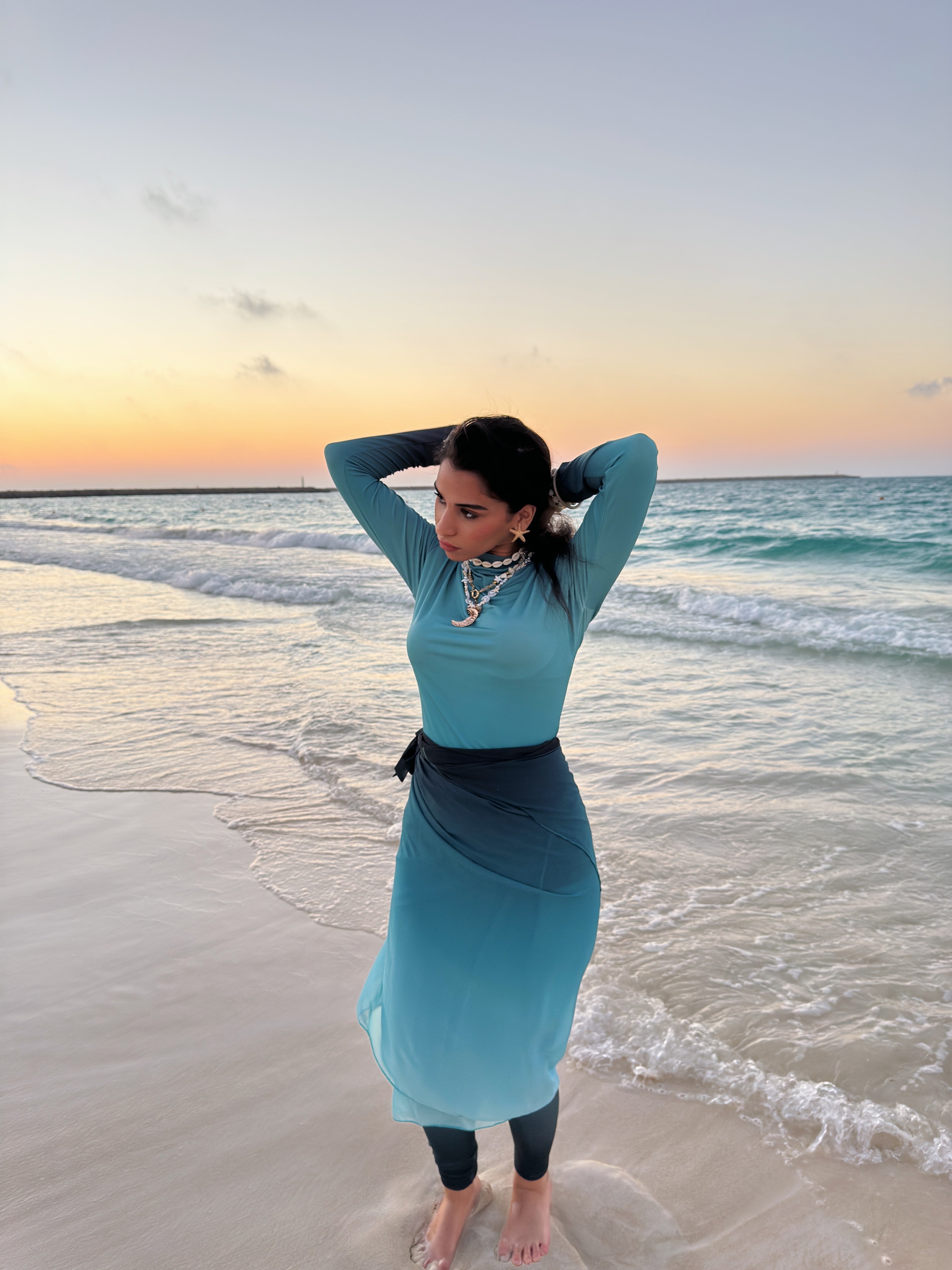 Ta’mayaa swimwear modest full coverage turquoise outfit worn by woman standing on beach at sunset