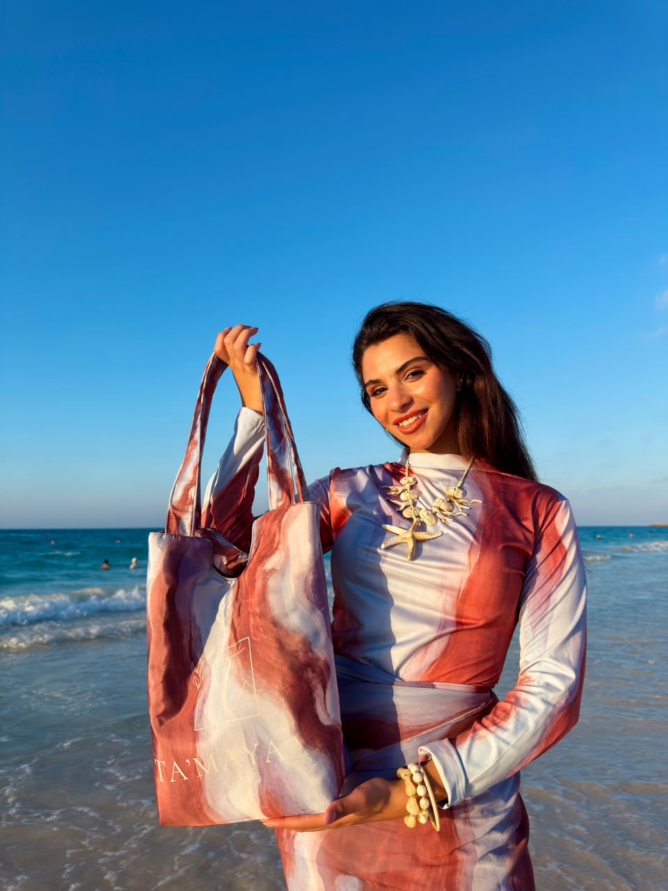 Woman holding colorful Ta’mayaa beach bag wearing stylish swimmers swimwear at the beach during sunset