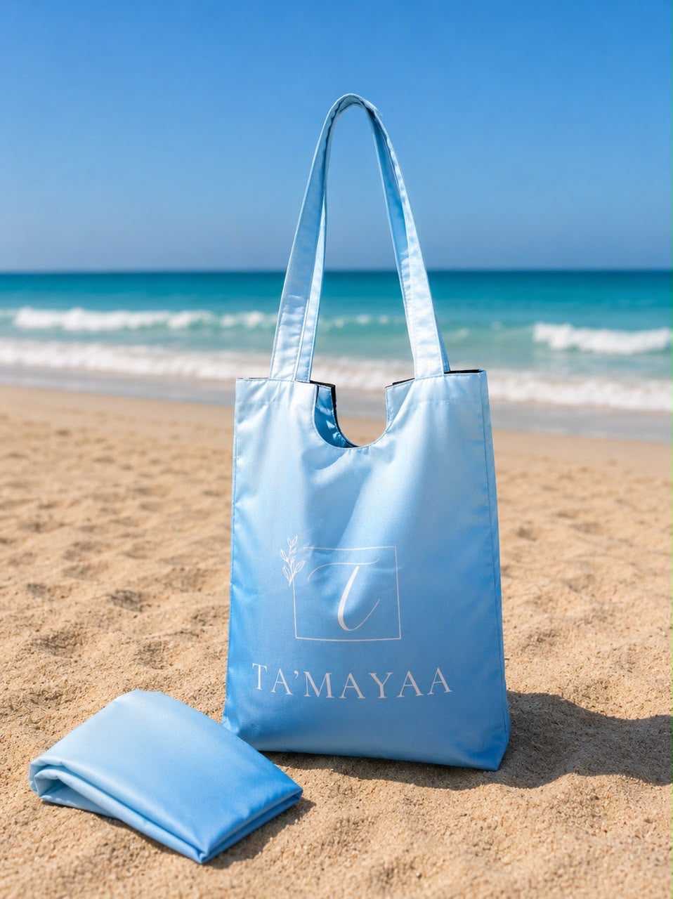 Light blue Ta’mayaa bag and swimwear modest folded on sandy beach with ocean background