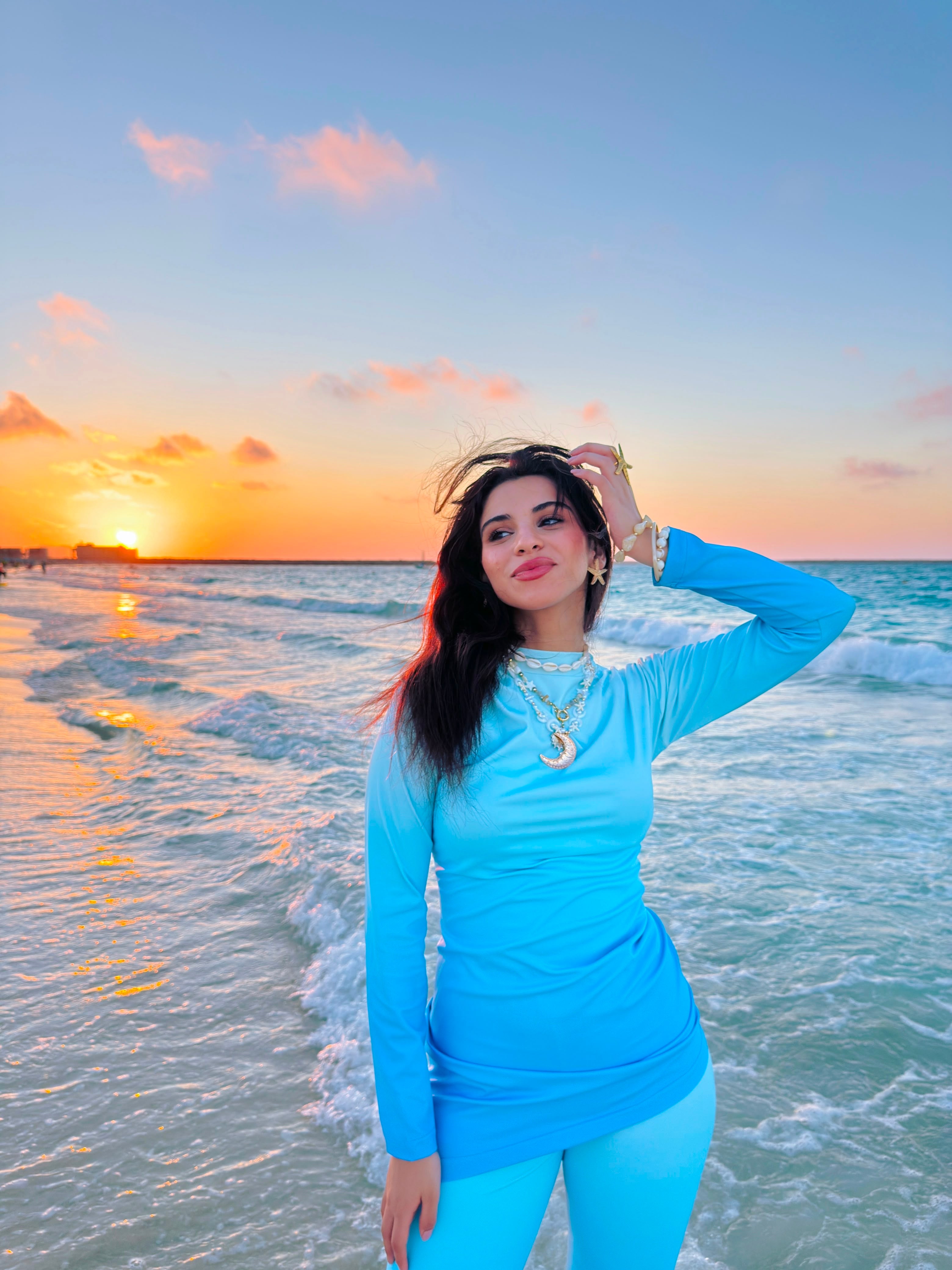Woman wearing Ta’mayaa swimwear modest in blue standing on the beach at sunset by the ocean
