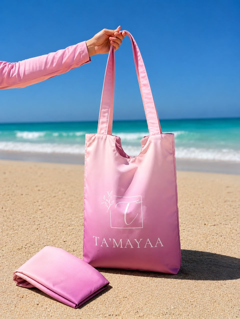 Pink Ta’mayaa swimwear modest bag and folded swimwear on a sandy beach with ocean background
